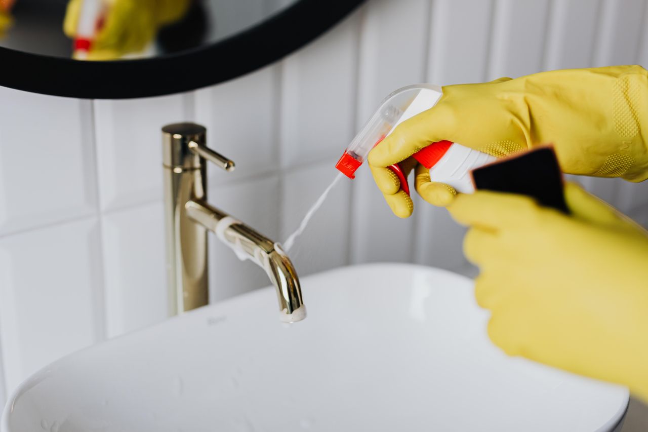 Seen from above, an anonymous housekeeper in yellow rubber gloves sprays cleaner onto a chrome bathroom faucet